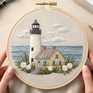 May include: Embroidered coastal scene in a wooden hoop. Features a white lighthouse with a black top, a yellow house with blue shutters, blue sea, white clouds, and flowers. The hoop is held by hands.