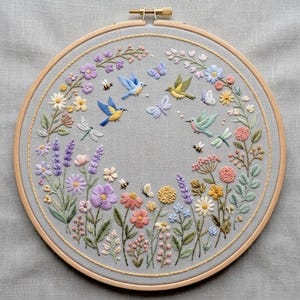 May include: A hand-stitched embroidery hoop featuring a floral and fauna design. The design includes colorful flowers, butterflies, bees, dragonflies, and birds in flight, all set against a gray fabric background. The wooden hoop is visible.