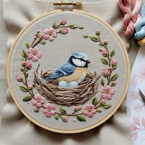 May include: Embroidered artwork featuring a blue bird in a nest with two eggs, surrounded by pink flowers and green leaves. The embroidery is set in a wooden hoop, creating a framed piece. The background is a neutral fabric.