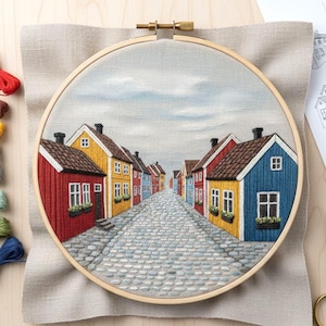 Peut inclure: Broderie sur tambour représentant une scène de rue avec des maisons colorées. Les maisons sont rouges, jaunes et bleues avec des toits bruns, sur fond de ciel gris. La rue pavée mène à l'horizon, créant une sensation de profondeur.