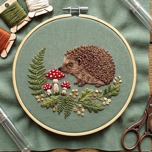 May include: An embroidery hoop containing a sage green fabric with a hand-stitched design of a brown hedgehog, three red and white spotted mushrooms, green fern leaves, and small yellow berries arranged in a circular pattern on the fabric.