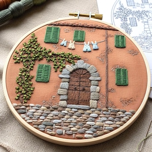 May include: Embroidered hoop art featuring a detailed depiction of a rustic building with a stone archway and wooden door. The artwork includes green shutters, a climbing vine, and miniature laundry hanging on a clothesline. The color palette includes shades of brown, green, and beige.