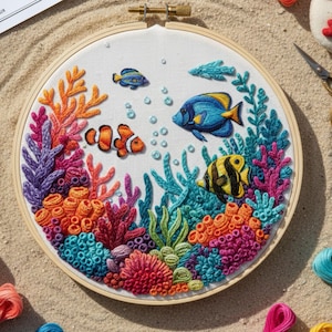 Puede incluir: Un bastidor de bordado colorido con una escena submarina. El diseño incluye arrecifes de coral en tonos naranja, rosa y morado, con peces azules y amarillos nadando entre el coral. El fondo es blanco y el bastidor es de madera.