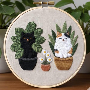 May include: Embroidered hoop art featuring two cats in pots with plants. One cat is black, the other is calico. A small pot of daisies is between them. The embroidery is on a beige fabric, set in a wooden hoop.