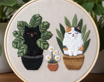 May include: Embroidered hoop art featuring two cats in pots with plants. One cat is black, the other is calico. A small pot of daisies is between them. The embroidery is on a beige fabric, set in a wooden hoop.
