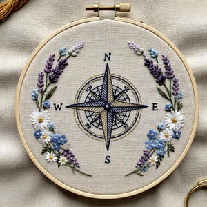 May include: Embroidered compass rose design in a wooden hoop. The compass is centered with a navy blue and gold star design, surrounded by floral embroidery in white, blue, and purple. The letters N, E, S, and W mark the cardinal directions.