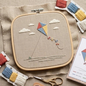 May include: Embroidered kite design in a square wooden hoop. The kite is blue, yellow, and red, with white clouds. Embroidery floss in various colors and a pair of gold scissors are also visible.