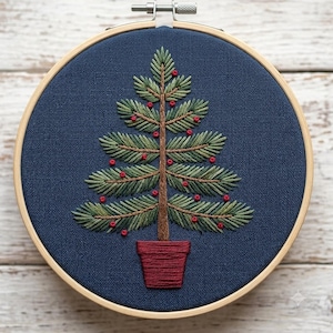 May include: A circular wooden embroidery hoop containing a navy blue fabric panel. The fabric features a hand-stitched design of a green pine tree with a brown trunk, decorated with small red knots, set inside a solid red embroidered flower pot.
