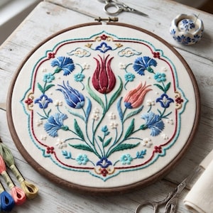 May include: A circular wooden embroidery hoop containing a white fabric panel with a hand-stitched floral design. The pattern features a central red tulip, a pink tulip, and various blue carnations and small white flowers, all framed by a red border.