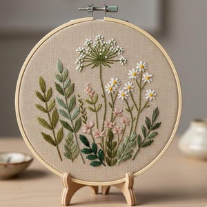 May include: A round embroidery hoop featuring a floral design. The embroidery showcases various wildflowers in shades of green, pink, and white, set against a beige fabric background. The hoop is held in a wooden stand.