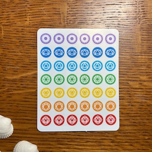 May include: A sheet of stickers with seven rows of colorful chakra symbols. Each row features a different color, starting with purple and ending with red. The symbols are white and outlined in black.