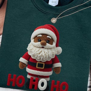 Peut inclure: Sweat-shirt vert foncé avec un motif de Père Noël au crochet. Le Père Noël porte un costume rouge et blanc avec une ceinture noire et une barbe blanche. Les mots "HO HO HO" sont brodés en rouge en bas.