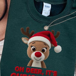 Peut inclure: Un sweat-shirt vert foncé avec un renne crocheté portant un bonnet de Père Noël rouge et blanc. Le renne a des bois bruns et un nez rouge. Le texte "OH DEER, IT'S CHRISTMAS" est brodé en rouge. Un collier argenté est également visible. Vêtements de Noël.