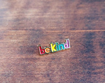 May include: A gold-colored enamel pin with the words "be kind" in rainbow colors. The pin is on a brown wooden surface.