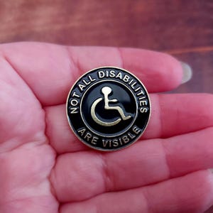 May include: A gold-colored pin with a black background. The pin features a wheelchair symbol and the text "Not all disabilities are visible".