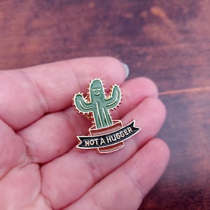 Puede incluir: Un pin de esmalte dorado con un cactus verde con una cara sonriente y el texto "NOT A HUGGER" en una pancarta negra.
