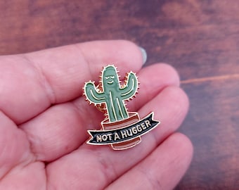May include: A gold-colored enamel pin featuring a green cactus with a smiling face and the text "NOT A HUGGER" on a black banner.