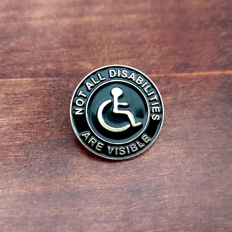 May include: A black and silver enamel pin with the text "Not All Disabilities Are Visible" and a wheelchair symbol in the center.