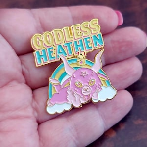 Puede incluir: Un pin de esmalte dorado con las palabras "GODLESS HEATHEN" en letras doradas y turquesas. El pin presenta un personaje de cabra rosa con una corona, acostado sobre un arcoíris y nubes.