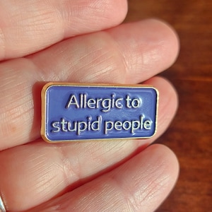 Può includere: Spilla rettangolare in smalto con bordo dorato e centro blu. La spilla recita "Allergic to stupid people" in lettere bianche. La spilla è tenuta in mano da una persona.