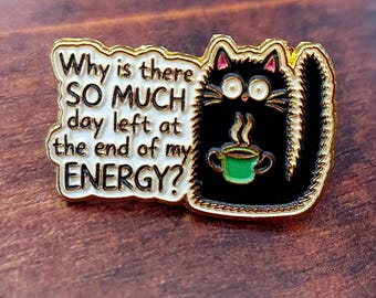 May include: A gold-rimmed enamel pin featuring a black cat with wide eyes holding a green coffee mug. The white speech bubble contains the black text: Why is there SO MUCH day left at the end of my ENERGY? The cat has pink inner ears.
