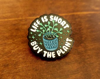 Puede incluir: Pin de esmalte negro con el texto "LIFE IS SHORT BUY THE PLANT" en letras blancas. El pin presenta una maceta azul con una planta verde en su interior. El pin es redondo con un borde festoneado.