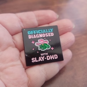 Puede incluir: Pin de esmalte negro con una rana verde que lleva un sombrero de vaquero rosa. El texto dice "Officially Diagnosed with Slay-DHD".