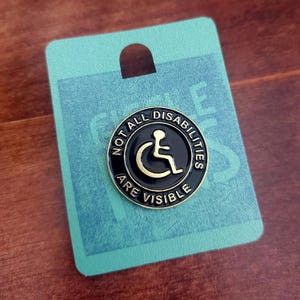 May include: A black and gold enamel pin with the text "Not all disabilities are visible" and a wheelchair symbol.