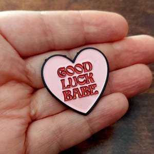 Puede incluir: Un pin de esmalte rosa en forma de corazón con las palabras "GOOD LUCK BABE" en letras rojas. El pin tiene un borde negro y es un accesorio lindo y positivo.