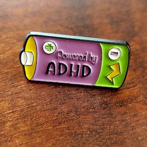 May include: A rectangular enamel pin shaped like a battery with a purple centre, yellow left end, and green right end. It features a white plus sign, a white minus sign, a yellow lightning bolt, and the black text Powered by ADHD.