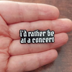 Könnte beinhalten: Eine schwarze Anstecknadel mit dem Text "i'd rather be at a concert" in weißer gotischer Schrift. Die Anstecknadel ist rechteckig und wird in der Hand einer Person gehalten.