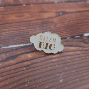 Puede incluir: Un pin de esmalte blanco con forma de nube con borde dorado. El pin presenta las palabras "DREAM BIG" en letras doradas. El pin está sobre un fondo de madera.