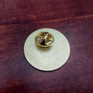 May include: The back of a gold-colored round pin with a pin back.