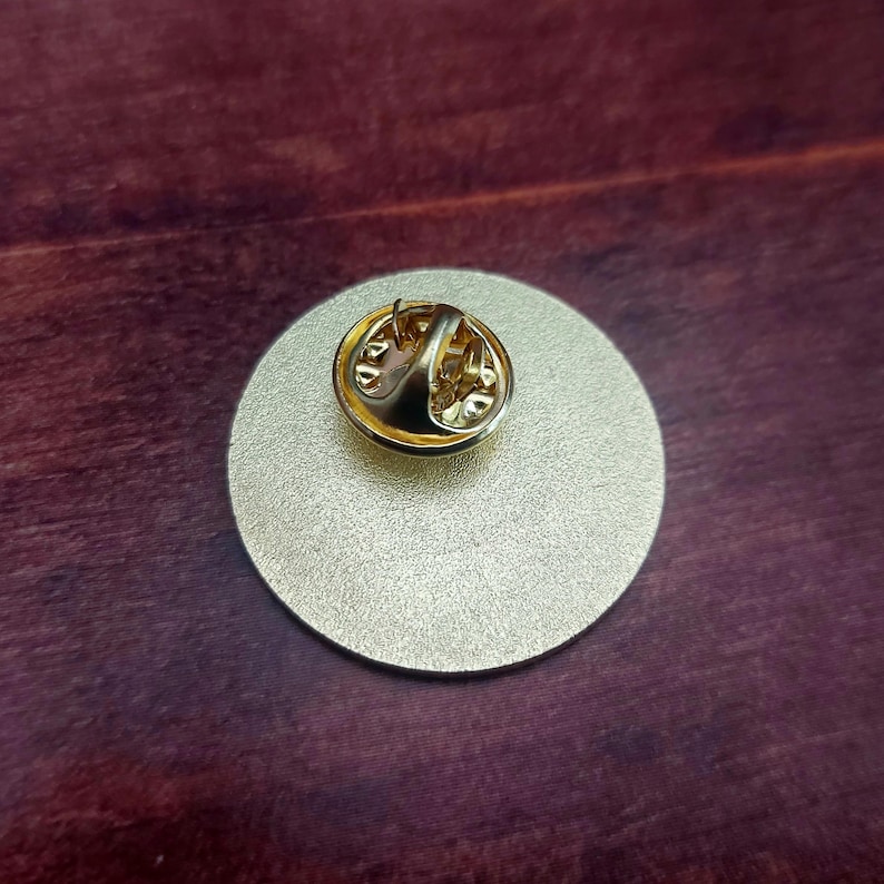 May include: The back of a gold-colored round pin with a pin back.