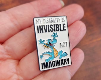 May include: A rectangular enamel pin with a black border and the text "MY DISABILITY IS INVISIBLE NOT IMAGINARY." A cartoon dragon in shades of blue and yellow is featured on the pin.