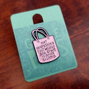 Op de afbeelding: Een roze emaille pin in de vorm van een draagtas met zwarte contouren. De pin heeft de tekst "NOT everything THAT WEIGHS you down IS yours to carry" in het wit. De pin zit op een teal kaart met een gat aan de bovenkant.