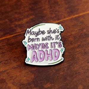 May include: An enamel pin with the text "Maybe she's born with it, maybe it's ADHD." The pin features a white background, purple text, and a black border. Green and blue accents are also present. The pin is on a wooden surface.