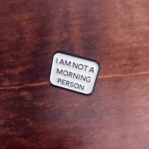 May include: White enamel pin with a black border. The pin reads "I am not a morning person".