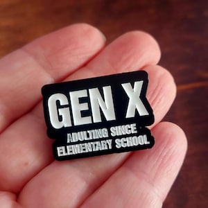 Puede incluir: Pin rectangular negro con las palabras "GEN X" en letras blancas. Debajo, el texto dice "ADULTING SINCE ELEMENTARY SCHOOL" en blanco. El pin se sostiene en una mano.