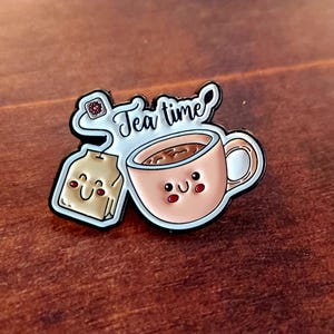 Peut inclure: Épingle en émail représentant une tasse de thé et un sachet de thé souriants, avec l'inscription "Tea time" dans une police fantaisiste. La tasse est de couleur pêche avec un intérieur marron, et le sachet de thé est beige.