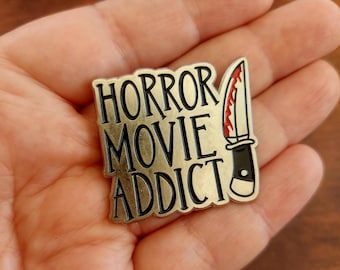 Puede incluir: Pin de esmalte dorado con las palabras "HORROR MOVIE ADDICT" en letras negras. Un cuchillo con una hoja roja está a la derecha. Un artículo de colección para los fanáticos del cine de terror.