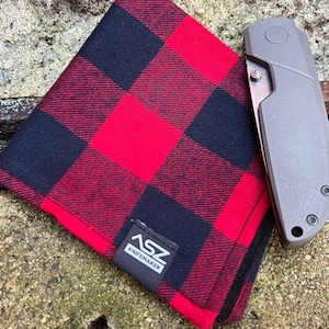 May include: A red and black checked fabric pocket square with a black leather patch that says "ASZ KNIFEMAKER". A grey pocket knife is lying on top of the fabric.