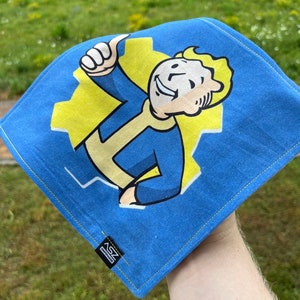 May include: Blue square cloth with a cartoon character giving a thumbs up, set against a yellow gear. The character is in blue and yellow. The cloth has a yellow stitched border and a small black tag with white text.
