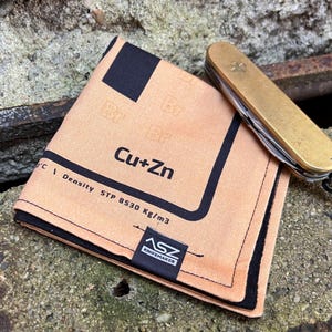 Puede incluir: Un pañuelo de tela marrón y negro con el texto "Cu+Zn Density STP 8530 kg/m3" y un logotipo negro que dice "ASZ Knifemaker".
