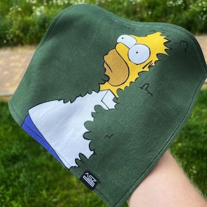 May include: A green square cloth with a cartoon depiction of Homer Simpson. The character's yellow face and hair are visible, along with his white shirt and blue collar. The cloth has a black border and the text "ASZ KNIFEMAKER" is printed on the bottom.