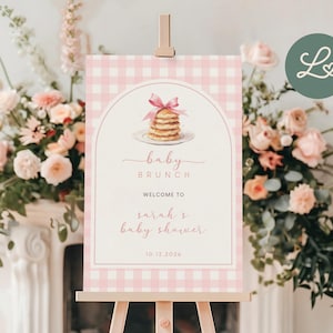 Puede incluir: Un letrero de baby brunch con un patrón de cuadros rosa y blanco, exhibido en un caballete de madera. El letrero incluye una ilustración de panqueques y el texto "baby brunch" y "welcome to sarah's baby shower" en una escritura elegante.