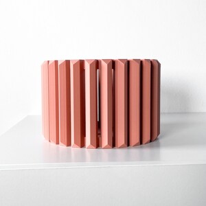 May include: A coral-colored, cylindrical decorative object with vertical, geometric slats. The object is placed on a white surface against a white background. The design is modern and minimalist, suitable for home decor.
