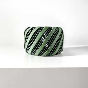 May include: A green, woven, cylindrical planter with a geometric pattern. The planter is made of a lightweight material and has a hollow center.