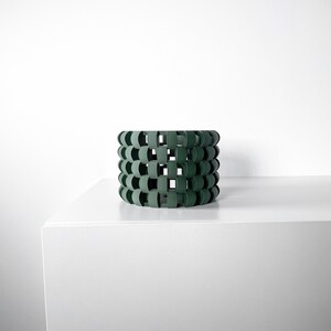 May include: A dark green cylindrical planter made of woven plastic. The planter has a geometric design with square and rounded shapes.