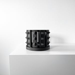 May include: A black, cylindrical decorative object with a series of vertical panels and conical spikes. The object sits on a circular base and is displayed against a white background. The spikes are evenly spaced around the circumference.